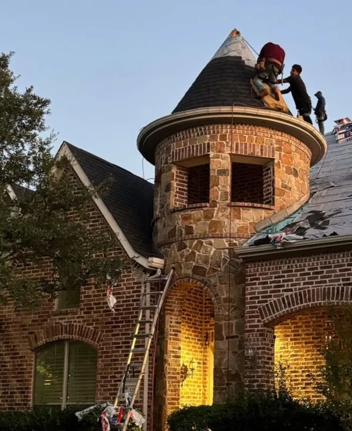 A crew shingling the turret of a brick and stone home at dusk